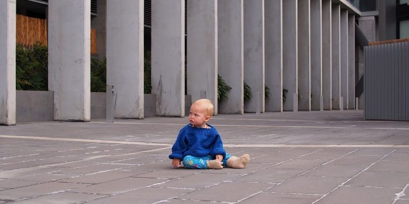 Young child about two and a half years old sitting on a featureless concrete yard at the base of a tower block